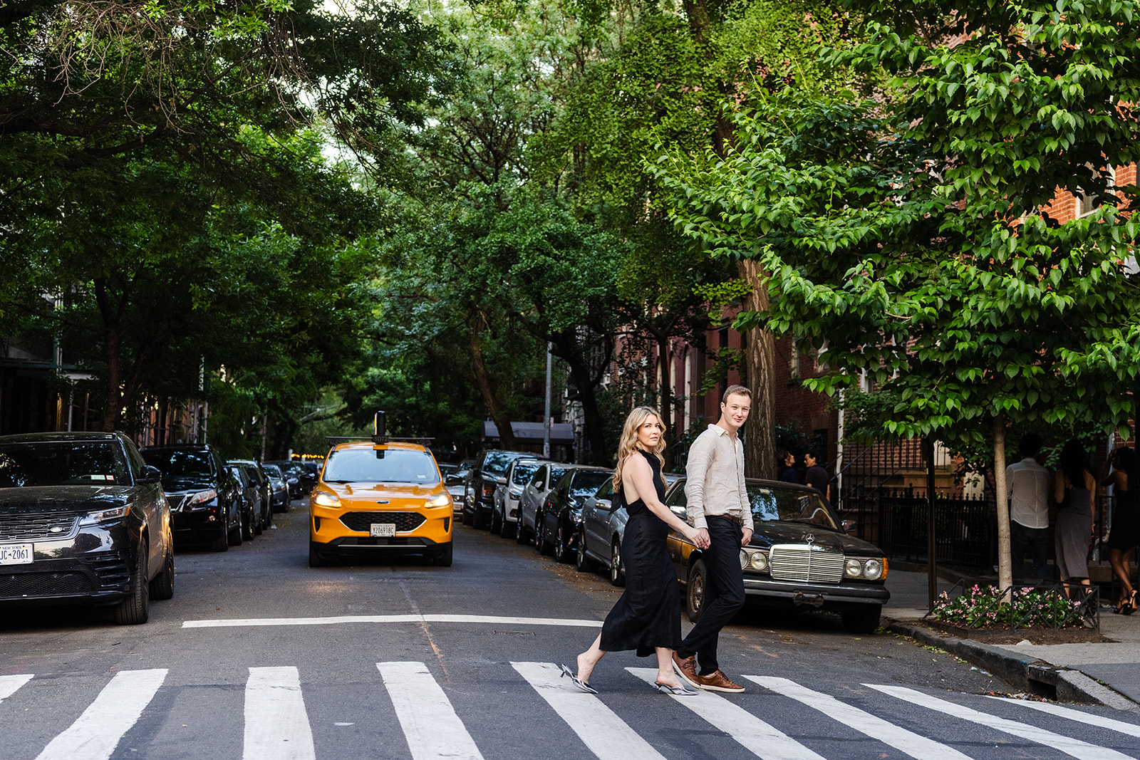 couple walking across a new york street, yellow cab behind them, green trees